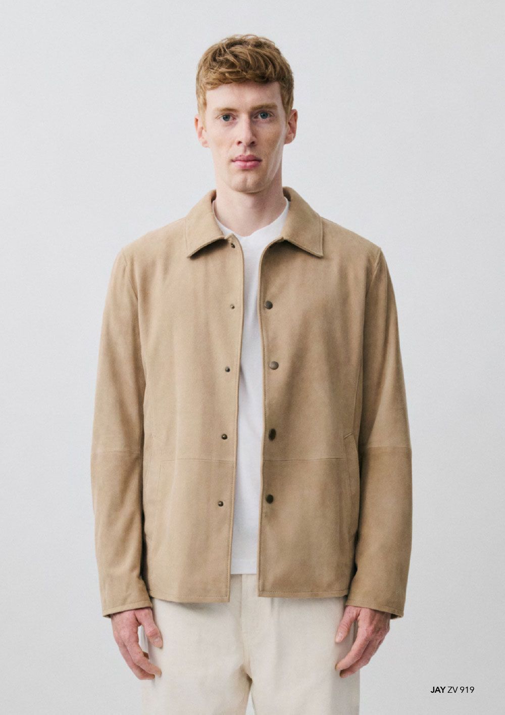 Eine Person, die eine hellbraune Wildlederjacke mit Knöpfen über einem weißen T-Shirt und einer hellen Hose trägt, vor einem neutralen Hintergrund.