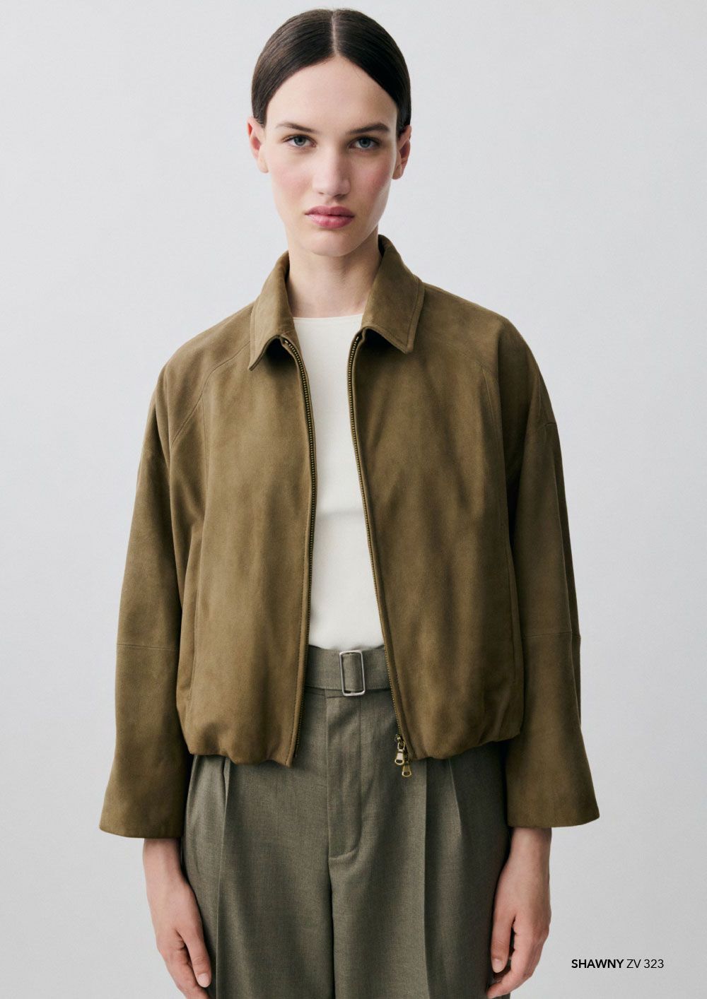 Ein Model posiert in einer grünen Wildlederjacke mit Reißverschluss über einem weißen Oberteil und einer grünen Hose vor einem schlichten hellen Hintergrund.