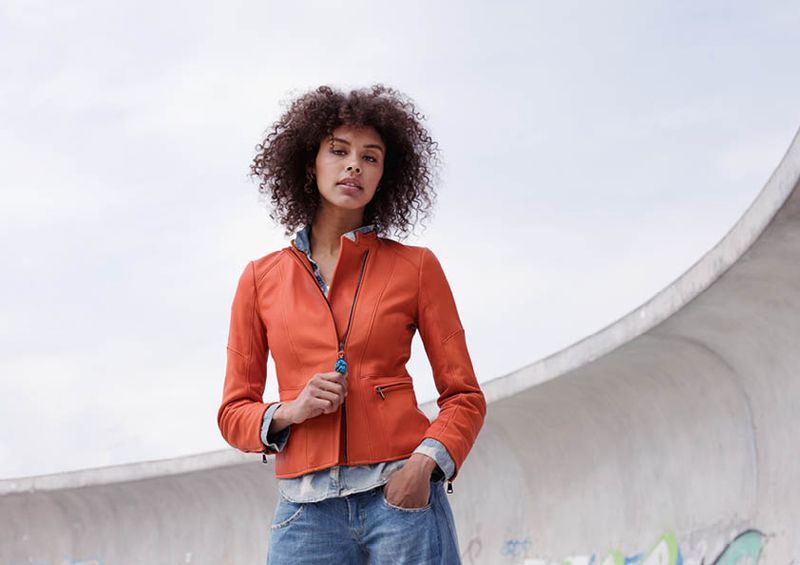 Eine Frau in einer orangefarbenen Lederjacke und Jeans steht vor einem Gebäude.