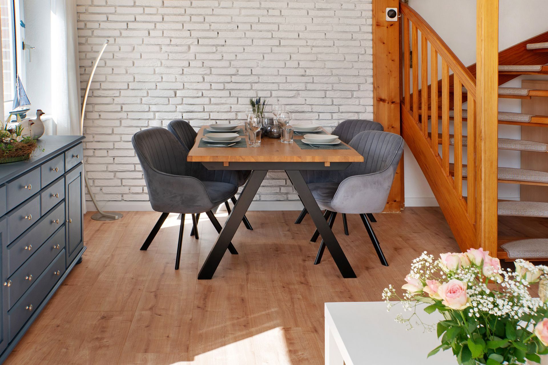 Esstisch mit vier Stühlen vor einer weißen Backsteinwand in der Nähe einer Treppe.