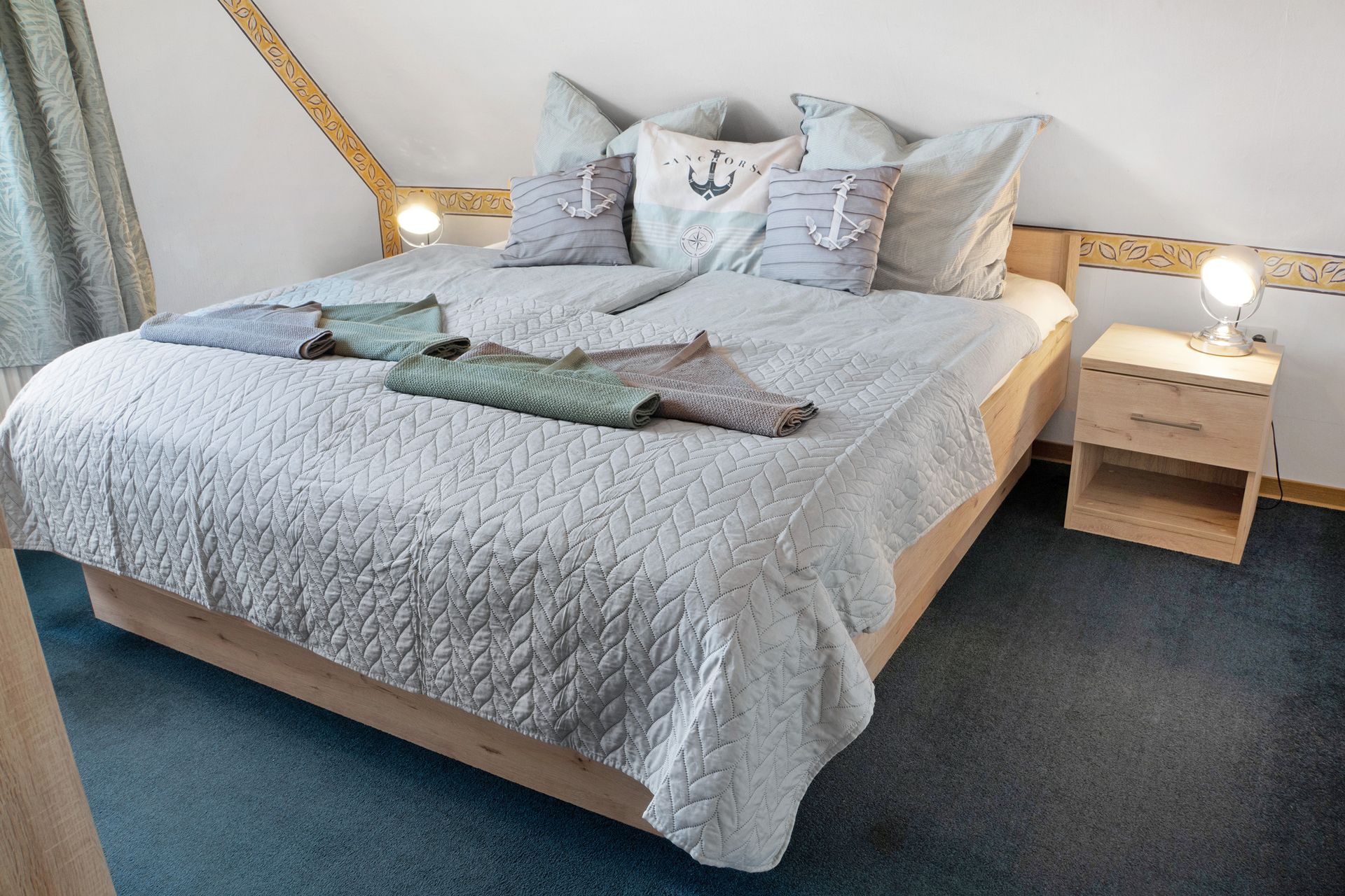 Schlafzimmer mit Bett, Kissen, Decke und zwei Nachttischen mit Lampen. Blauer Teppich.