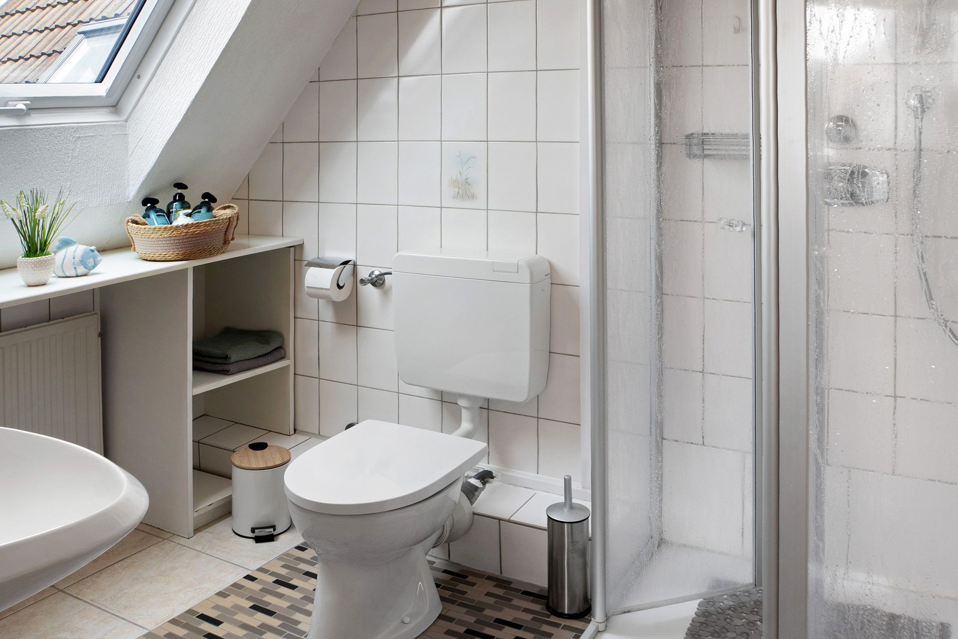 Weißes Badezimmer mit Toilette, Dusche und Waschbecken unter einer Dachschräge mit Fenster.