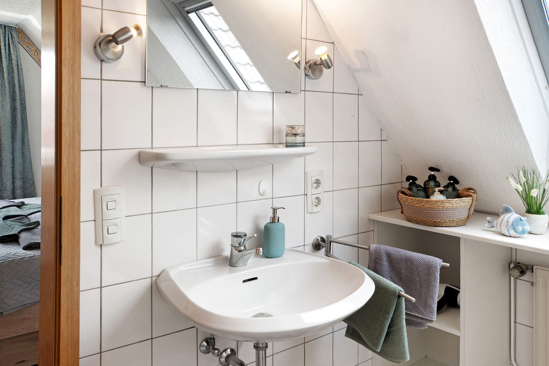 Badezimmer mit weißem Waschbecken, Spiegel und Ablage; schräge Wand mit Oberlicht.