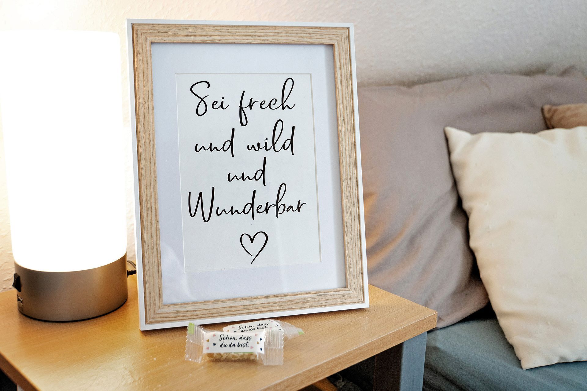 Ein gerahmtes Kalligrafiebild mit einem Zitat in deutscher Sprache, neben einer Lampe und einem Sofa.