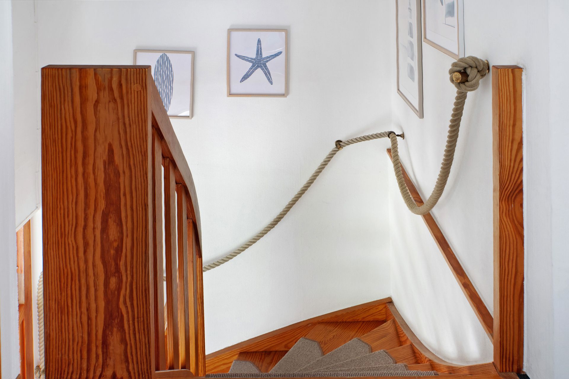 Treppe mit Holzgeländer, Seilhandlauf, nautischer Dekoration: gerahmte Kunstwerke und ein Seilknoten an der Wand.