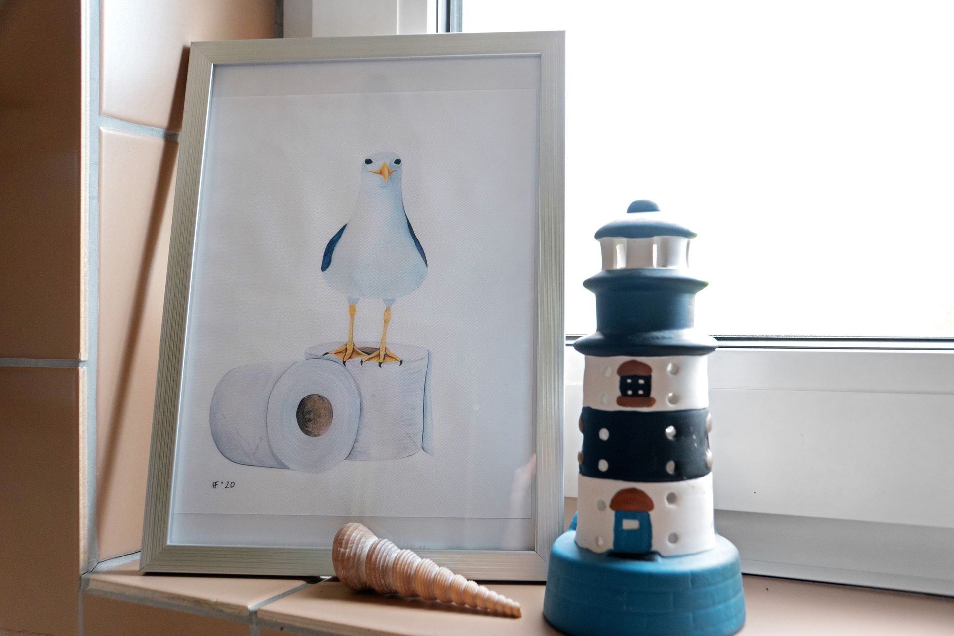 Möwe auf Toilettenpapier, gerahmter Druck, neben Leuchtturm und Muschel, auf einem Fensterbrett.