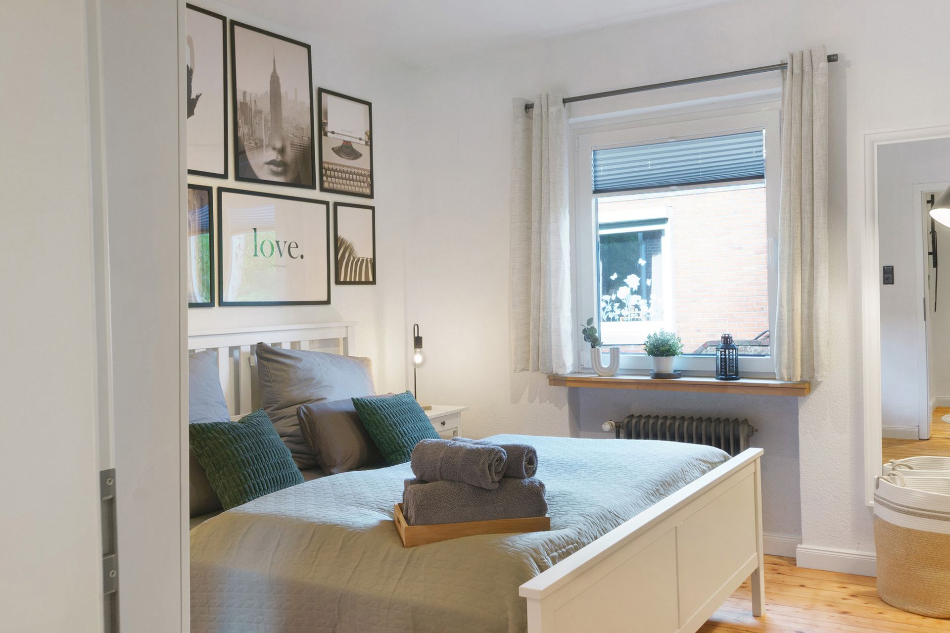 Gemütliches Schlafzimmer mit weißen Wänden, gerahmten Bildern und einem Bett mit Kissen und Handtüchern. Fenster mit Vorhängen und Heizkörper.