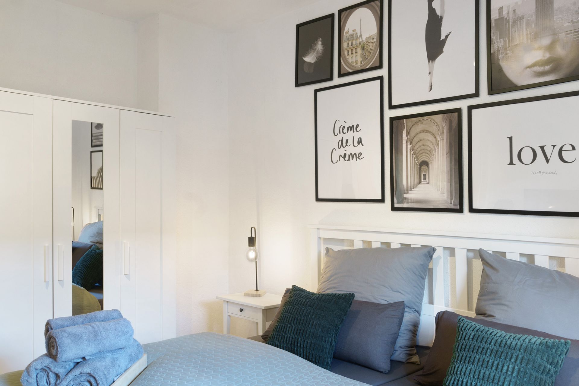 Schlafzimmer mit weißem Bett, grauen Kissen, gerahmten Bildern und einem weißen Kleiderschrank mit Spiegel.