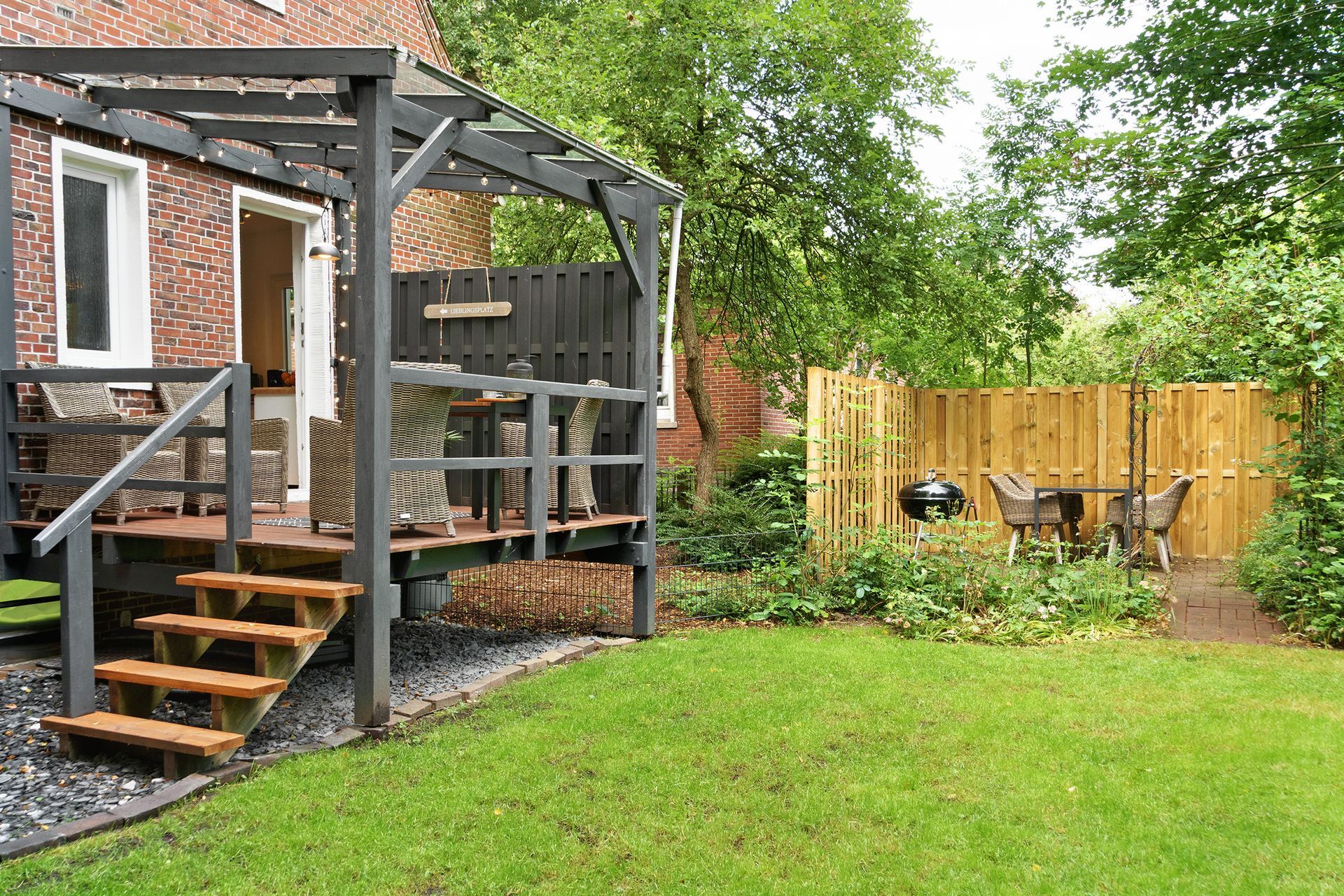 Holzterrasse mit Pergola, Treppe und Garten mit Grill neben einem Holzzaun.