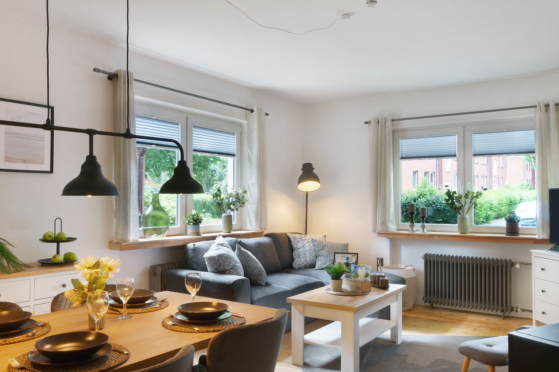 Wohnzimmer mit grauem Sofa, kleinem weißen Couchtisch und großen Fenstern, die viel Tageslicht hereinlassen.