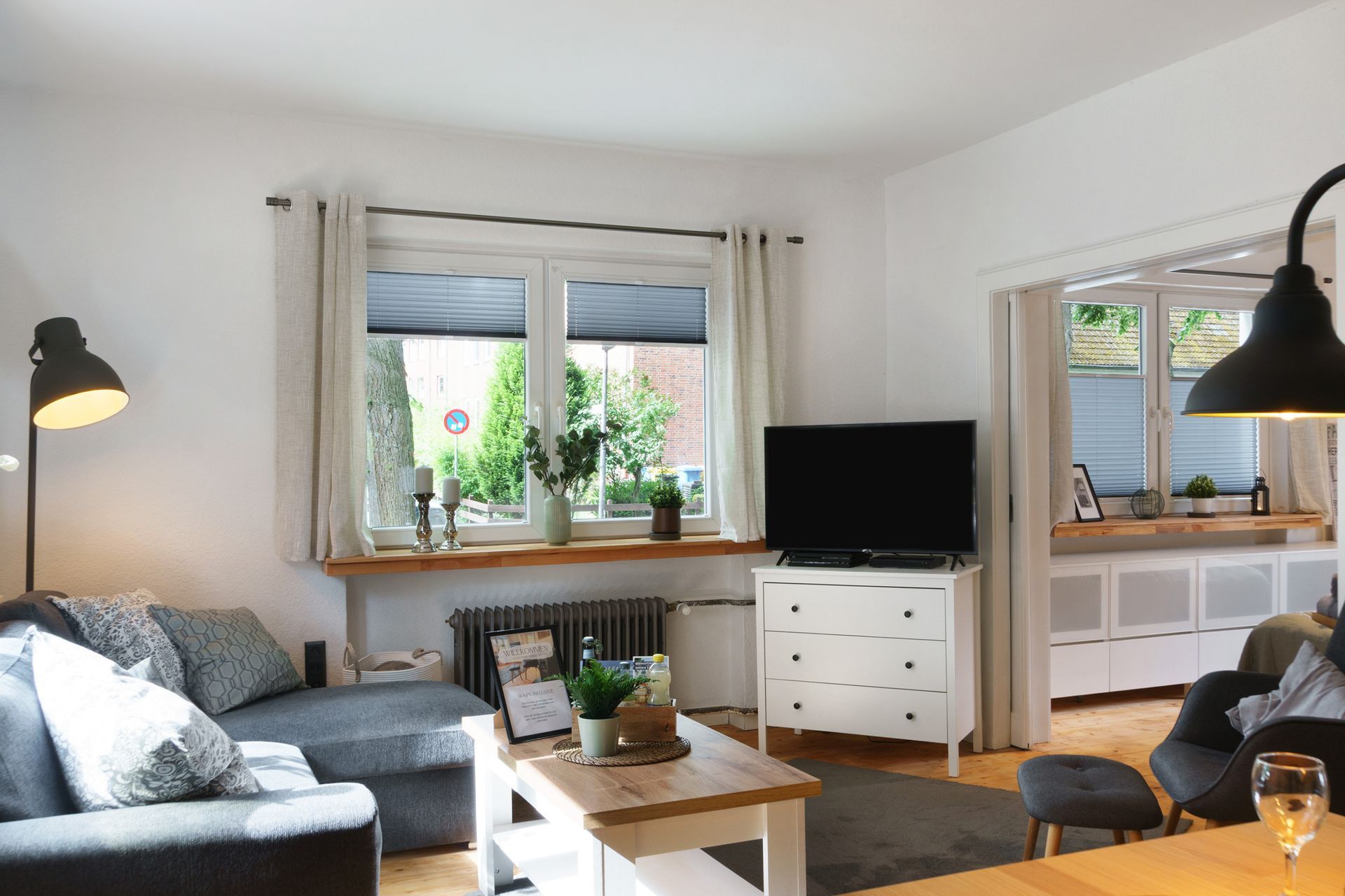 Wohnzimmer mit Sofa, Fernseher auf einer weißen Kommode und Fenster mit Blick ins Grüne.