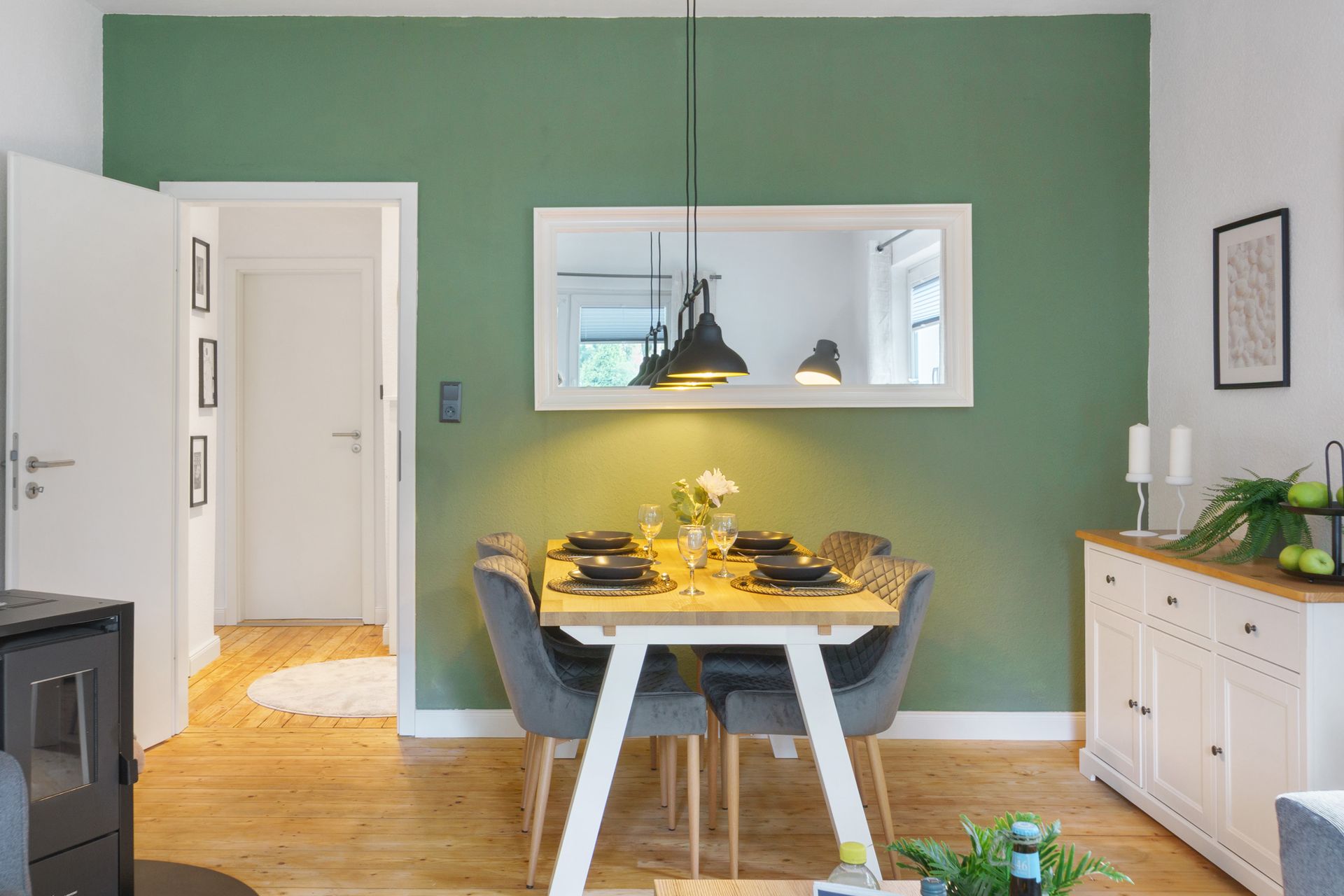 Esszimmer mit grüner Wand, rechteckigem Spiegel, Holztisch, grauen Stühlen und weißem Sideboard.