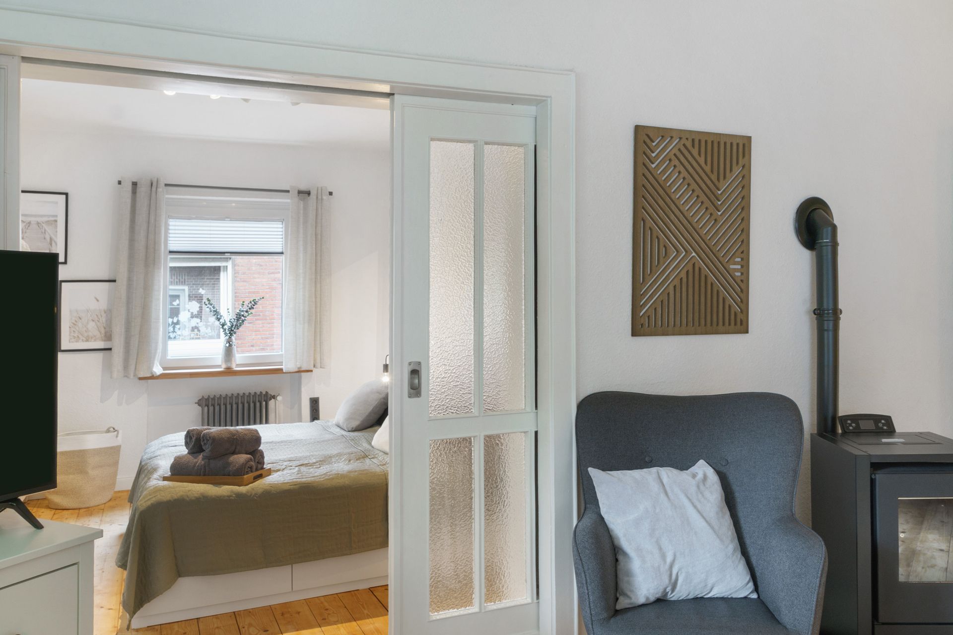 Schlafzimmerinterieur mit Bett, Sessel, Kunstwerken und teilweise geöffneter Milchglastür.