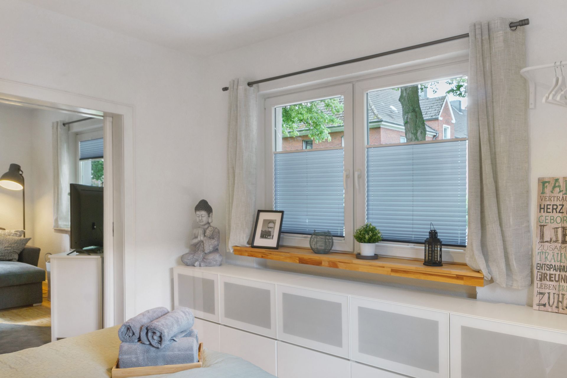 Helles Schlafzimmerinterieur mit Fenster, Holzregal, weißen Schränken und Buddha-Statue.