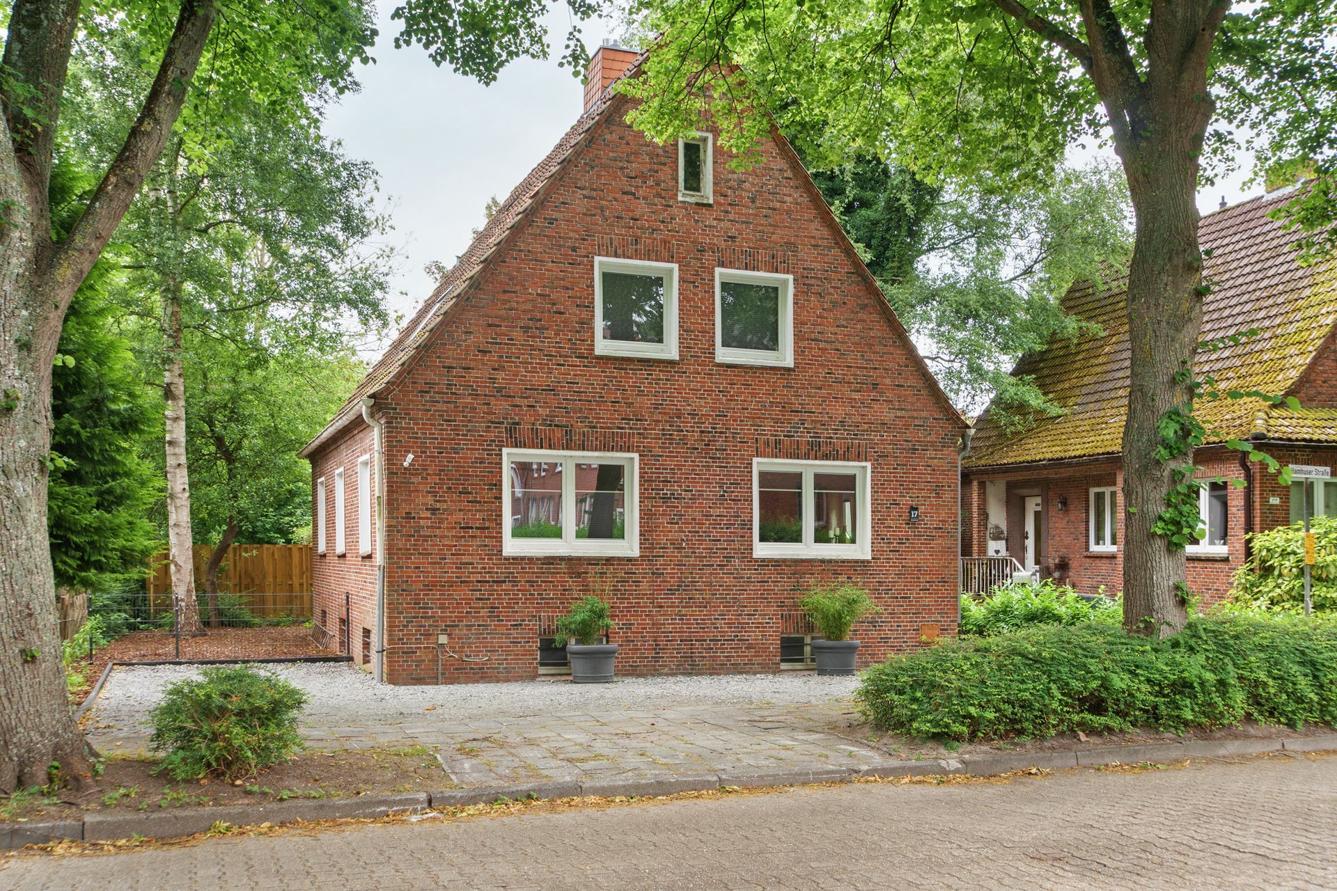Backsteinhaus mit weißgerahmten Fenstern, umgeben von Bäumen und Grün.