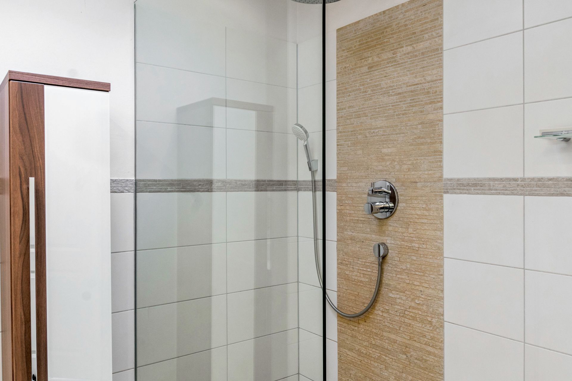 Dusche mit Glastür, beige Mosaikfliesenwand, weiß gefliester Wand, Chromarmaturen und einem Holzschrank.