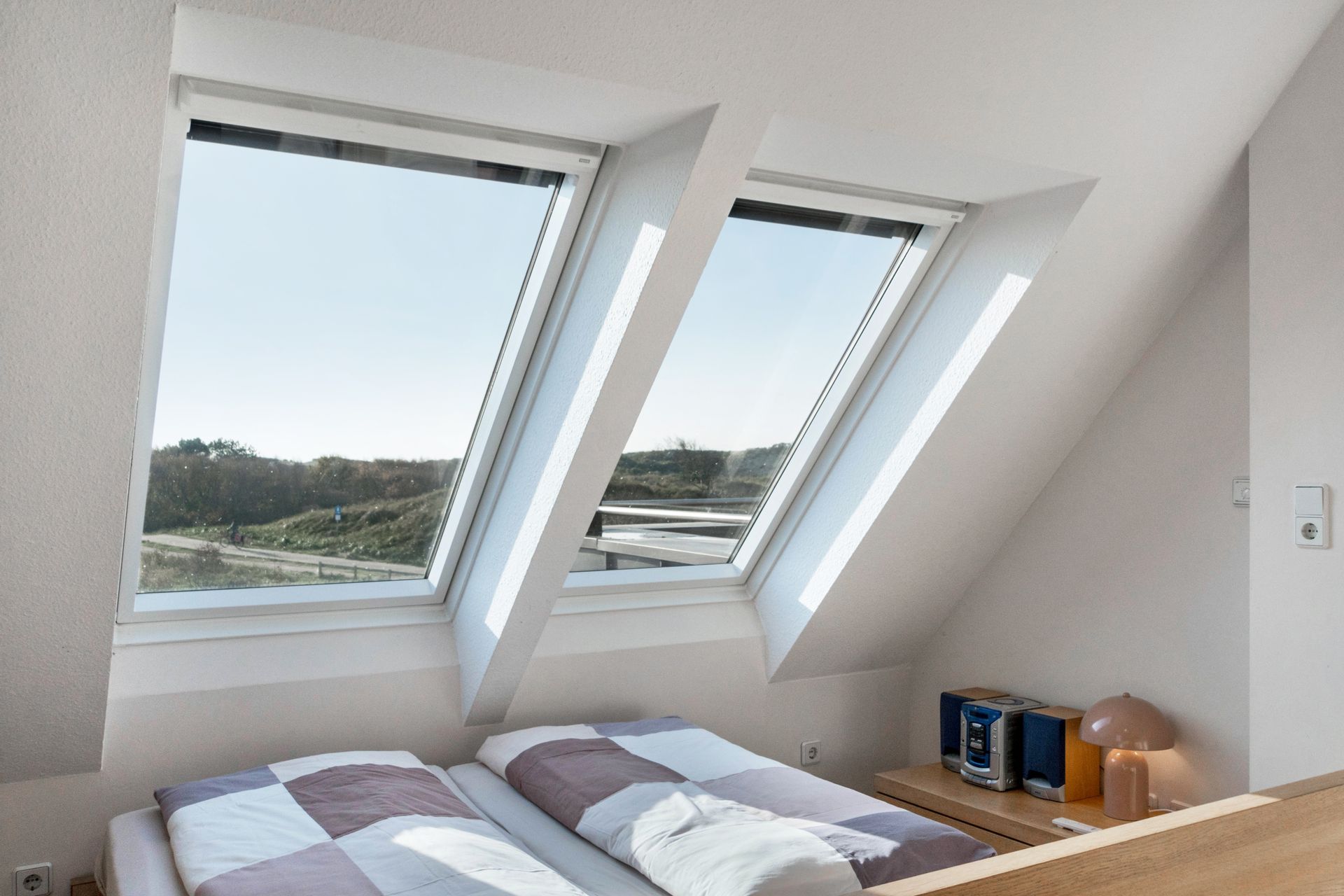 Zwei schräge Dachfenster in einem Dachgeschossschlafzimmer mit Blick auf die Landschaft. Ein Bett und ein Nachttisch sind zu sehen.