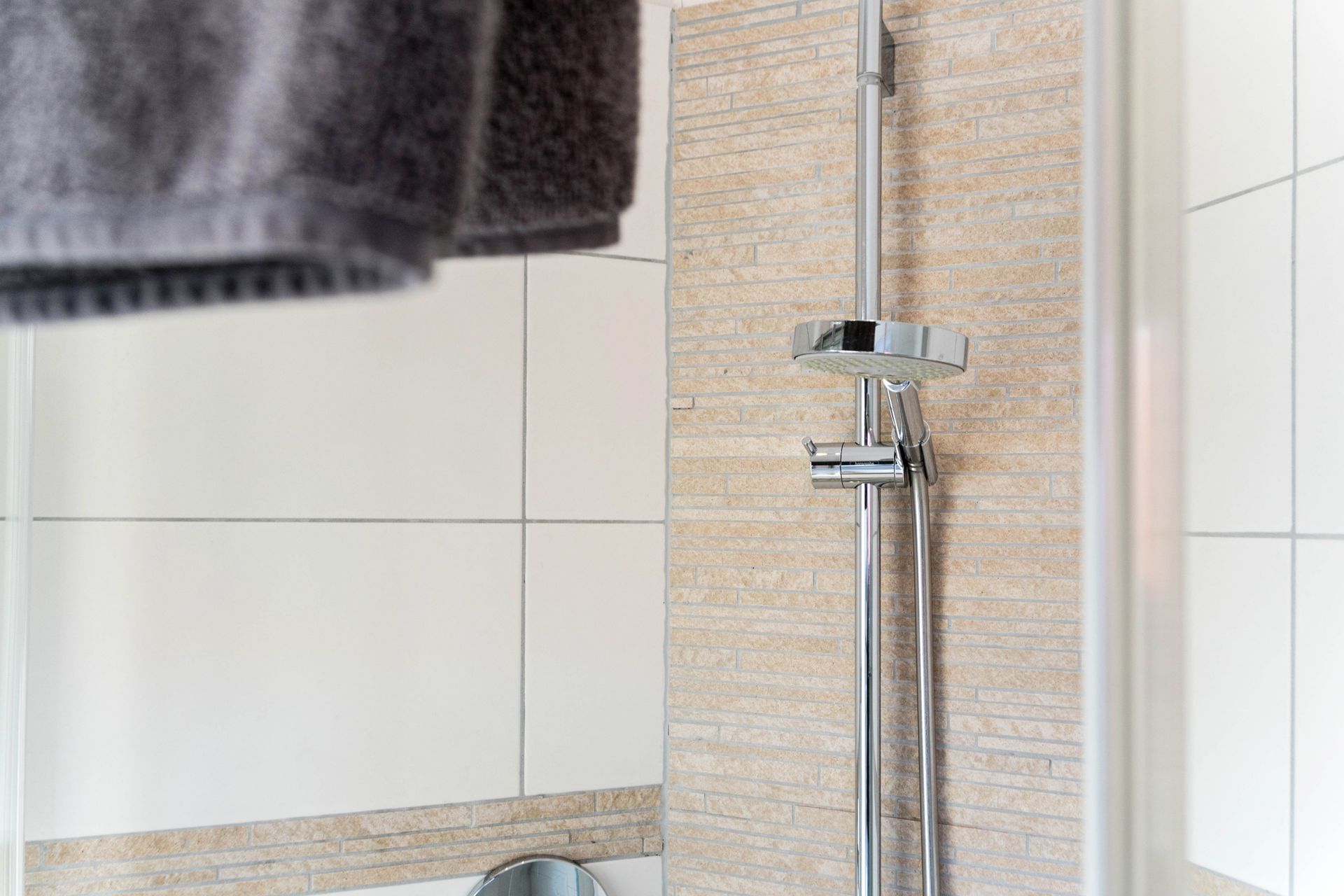 Duschkopf und Haltestange vor strukturierter Wand, weiße Fliesen und ein Handtuch.