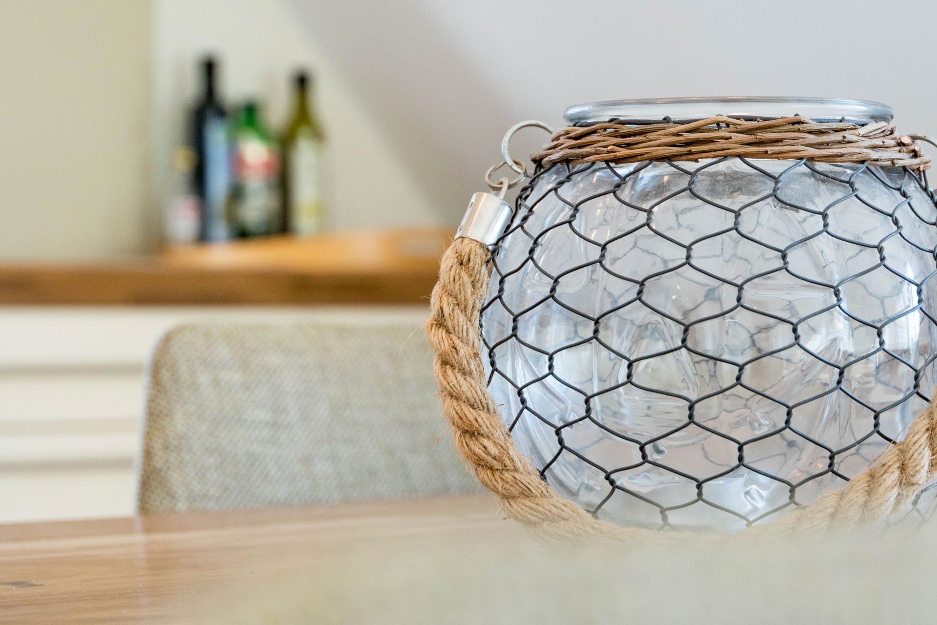 Ein mit Hühnerdraht, Seil und Weide umwickeltes Glasgefäß steht auf einem Holztisch. Flaschen sind im Hintergrund verschwommen zu sehen.