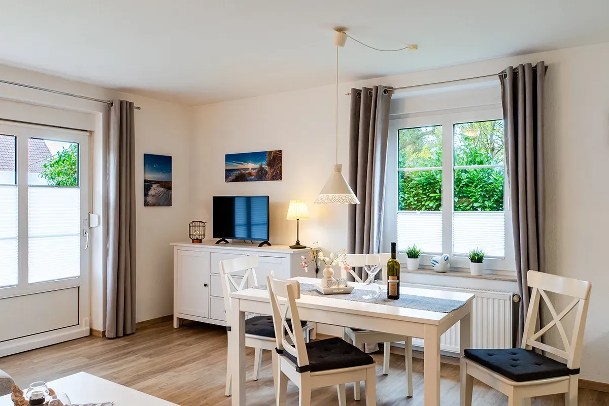 Wohnzimmer mit weißen Möbeln: Esstisch, Stühle, Sideboard, Fernseher. Fenster mit grauen Vorhängen, Tür nach draußen.