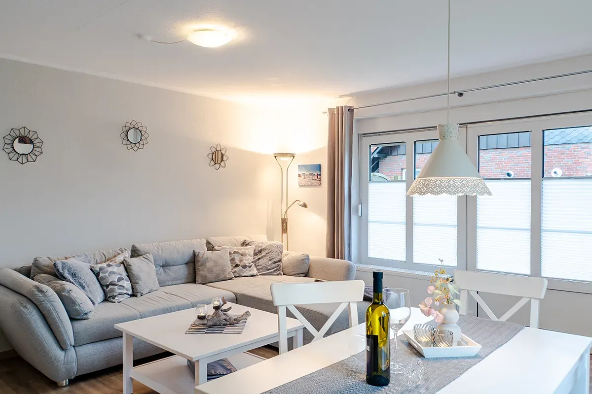 Wohnzimmer mit grauem Ecksofa, weißem Tisch, Esstisch und großem Fenster.