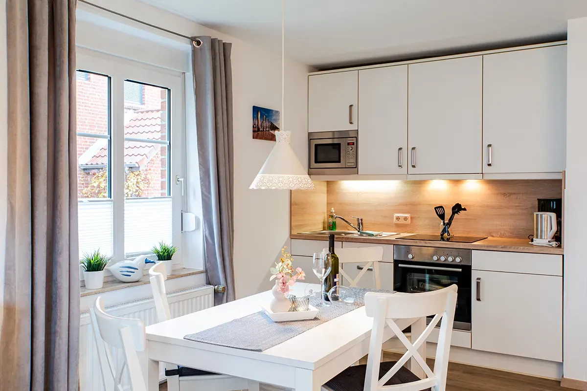 Küche mit weißen Schränken, gedecktem Esstisch und Fenster mit Vorhängen.