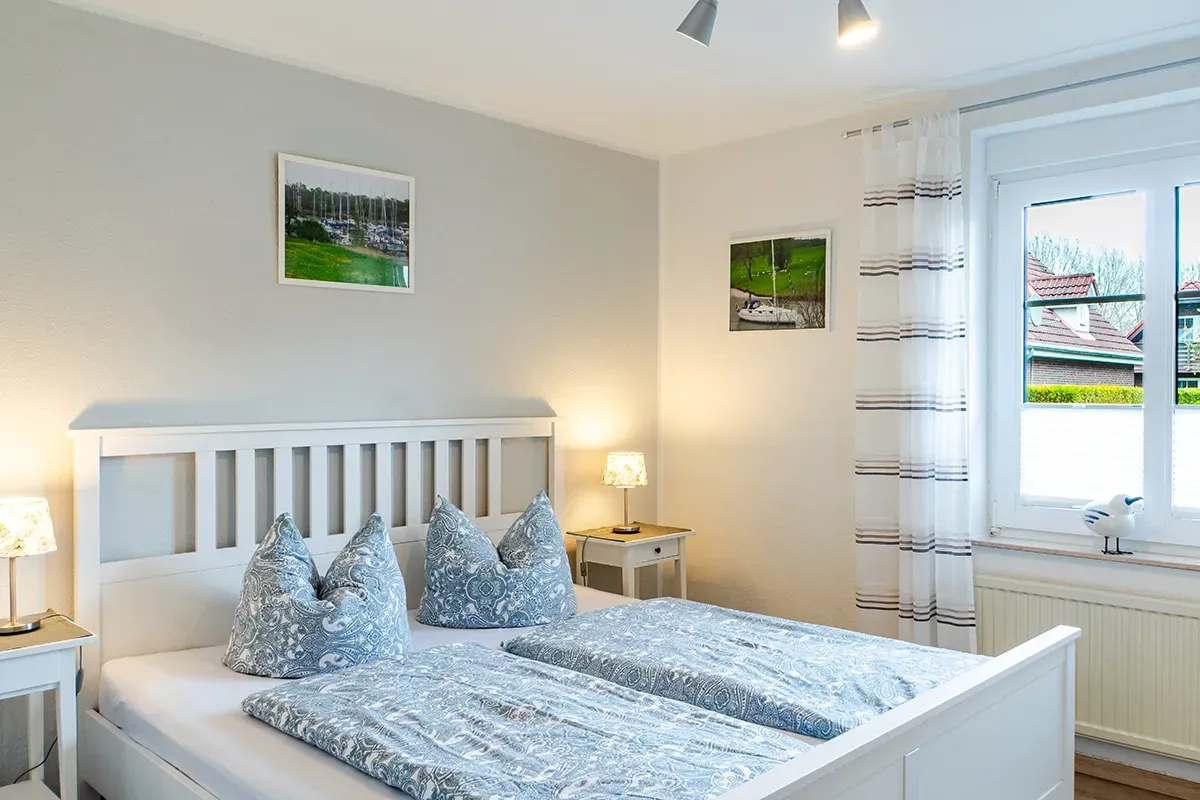 Schlafzimmer mit weißem Bett, grauer Bettwäsche, Nachttischen und einem Fenster mit Aussicht.