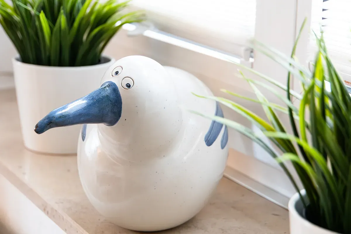 Weißer Keramikvogel mit blauem Schnabel und blauen Akzenten, auf der Fensterbank neben Topfpflanzen.