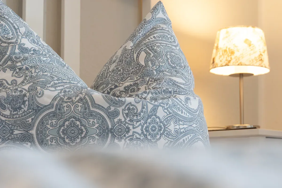 Zwei gemusterte Kissen auf einem Bett, daneben ein beleuchteter Lampenschirm.
