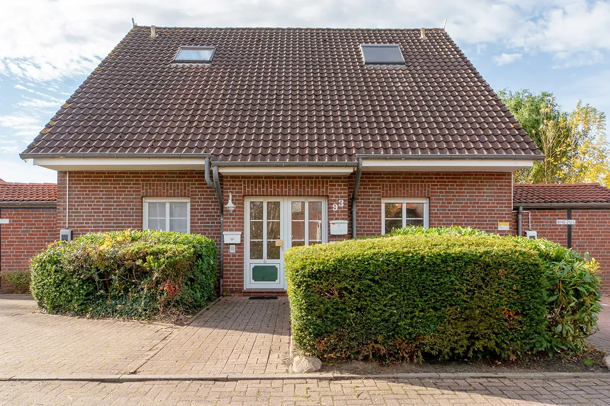 Backsteinhaus mit braunem Ziegeldach, zwei Fenstern und grüner Hecke.