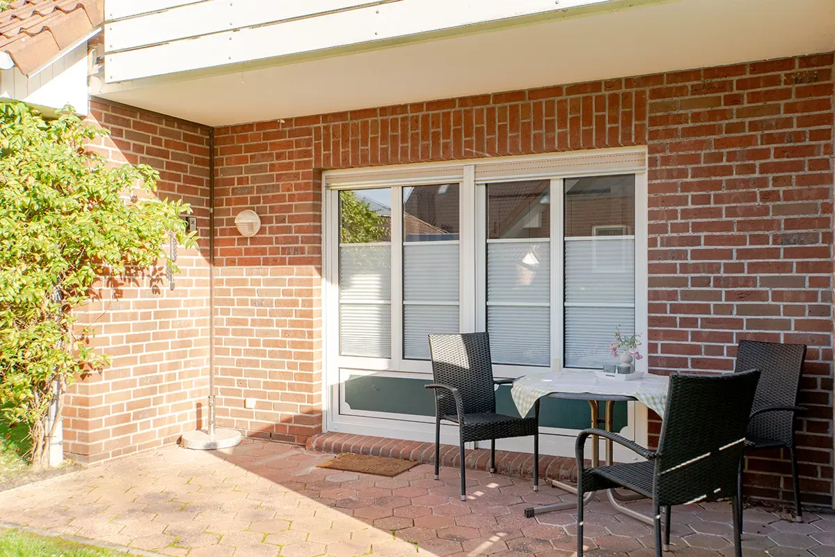 Eine kleine Terrasse mit Tisch und Stühlen vor einem Backsteingebäude mit weiß gerahmten Fenstern.