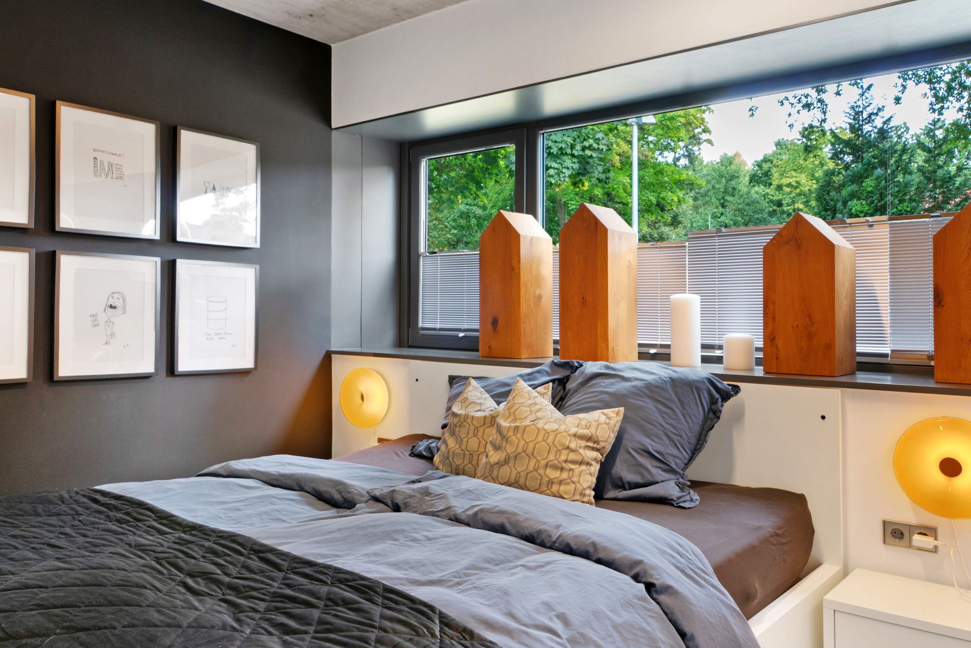 Schlafzimmer mit grauer Bettwäsche, Kunstwerken an der Wand und Holzelementen an einem Fenster mit Blick ins Grüne.