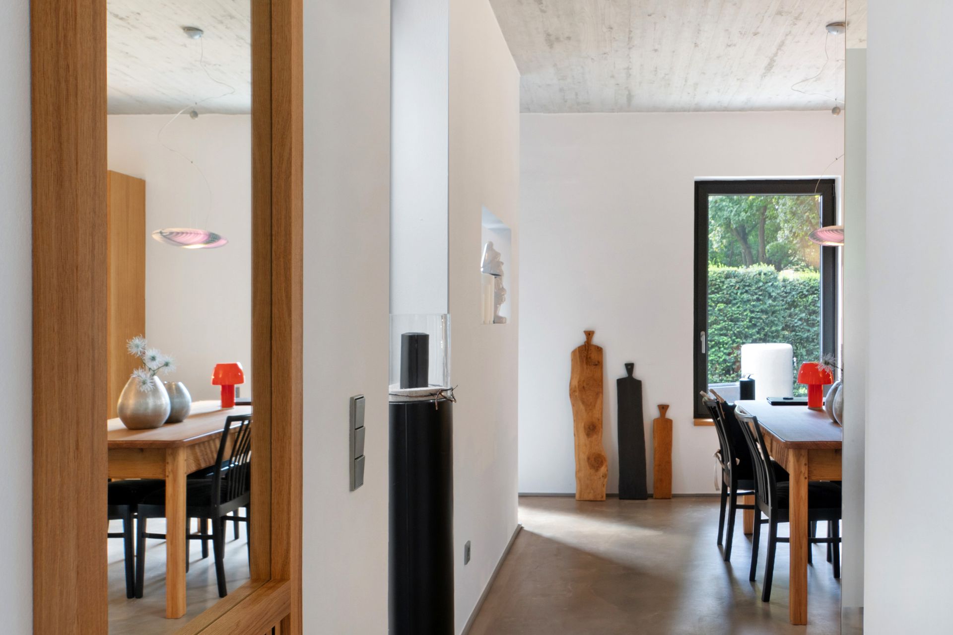 Flur mit Blick auf den Essbereich; hölzerner Türrahmen, weiße Wände, Betondecke, Tische, schwarze Stühle, Kunstwerke und Fenster.