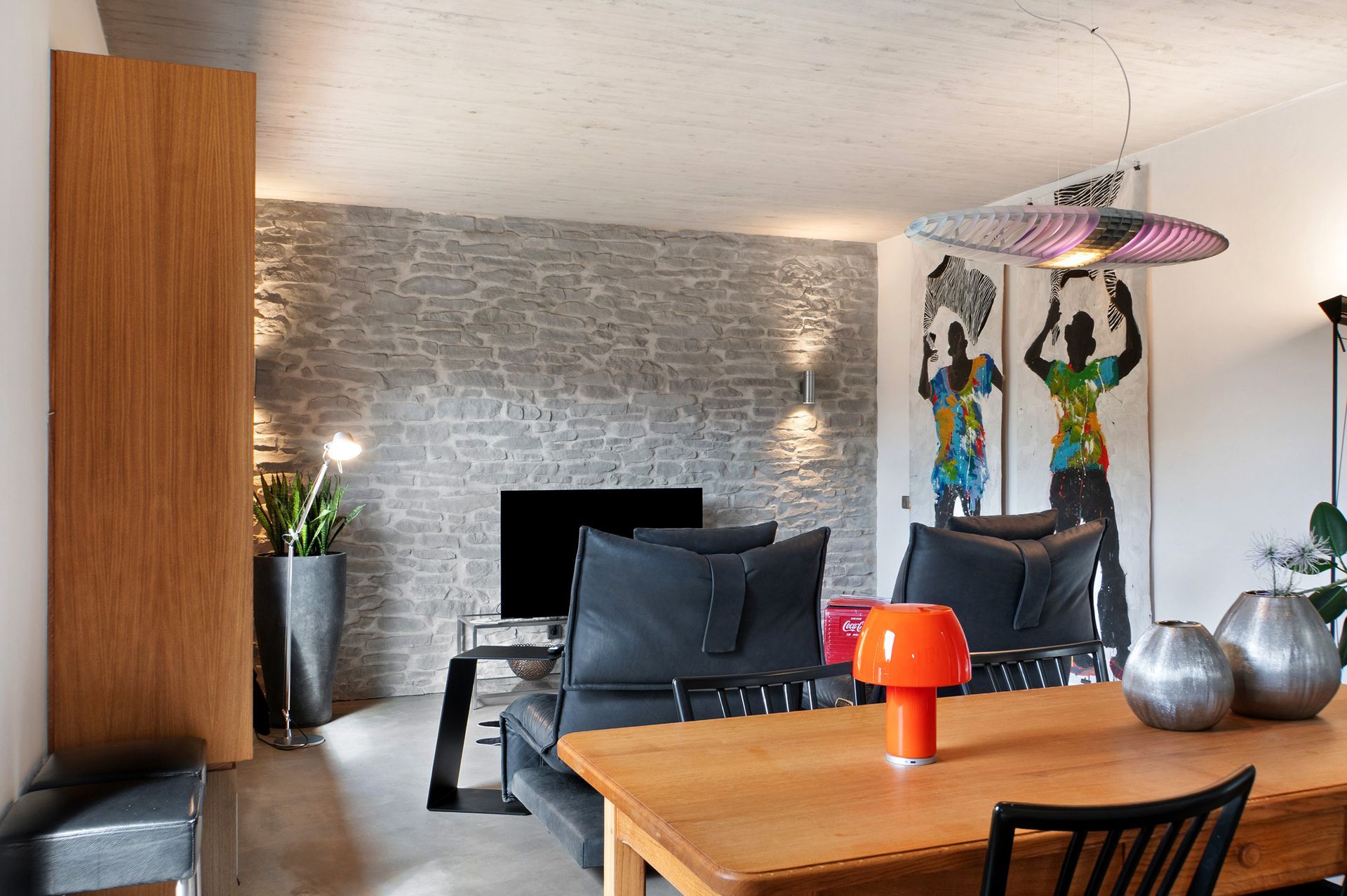 Wohnzimmer mit Steinwand, Fernseher und schwarzem Ledersofa. Holztisch mit orangefarbener Lampe und Kunstwerken.
