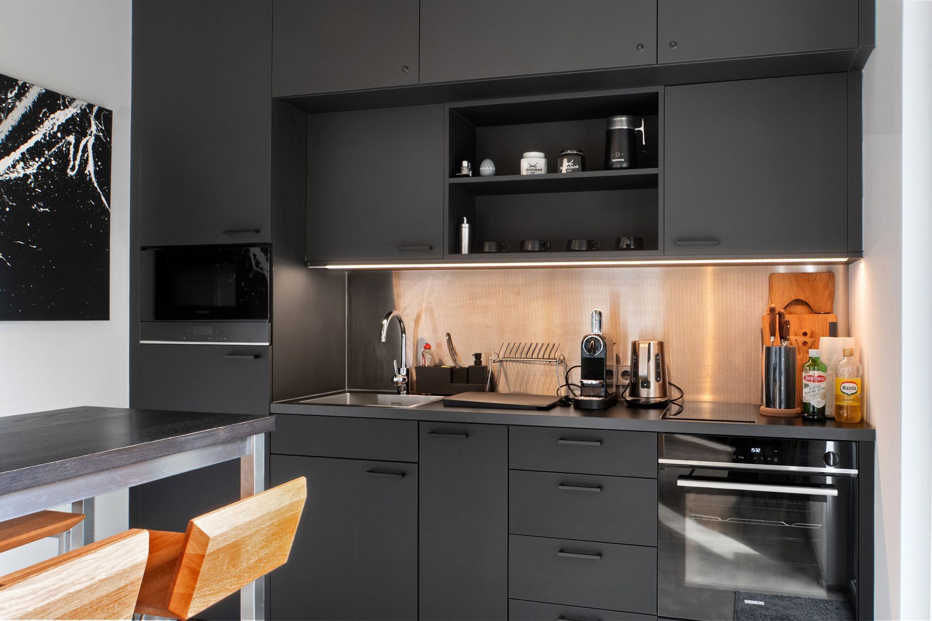 Dunkelgraue, moderne Küche mit Edelstahl-Spritzschutz, Einbaugeräten und einem kleinen Tisch mit Stühlen.