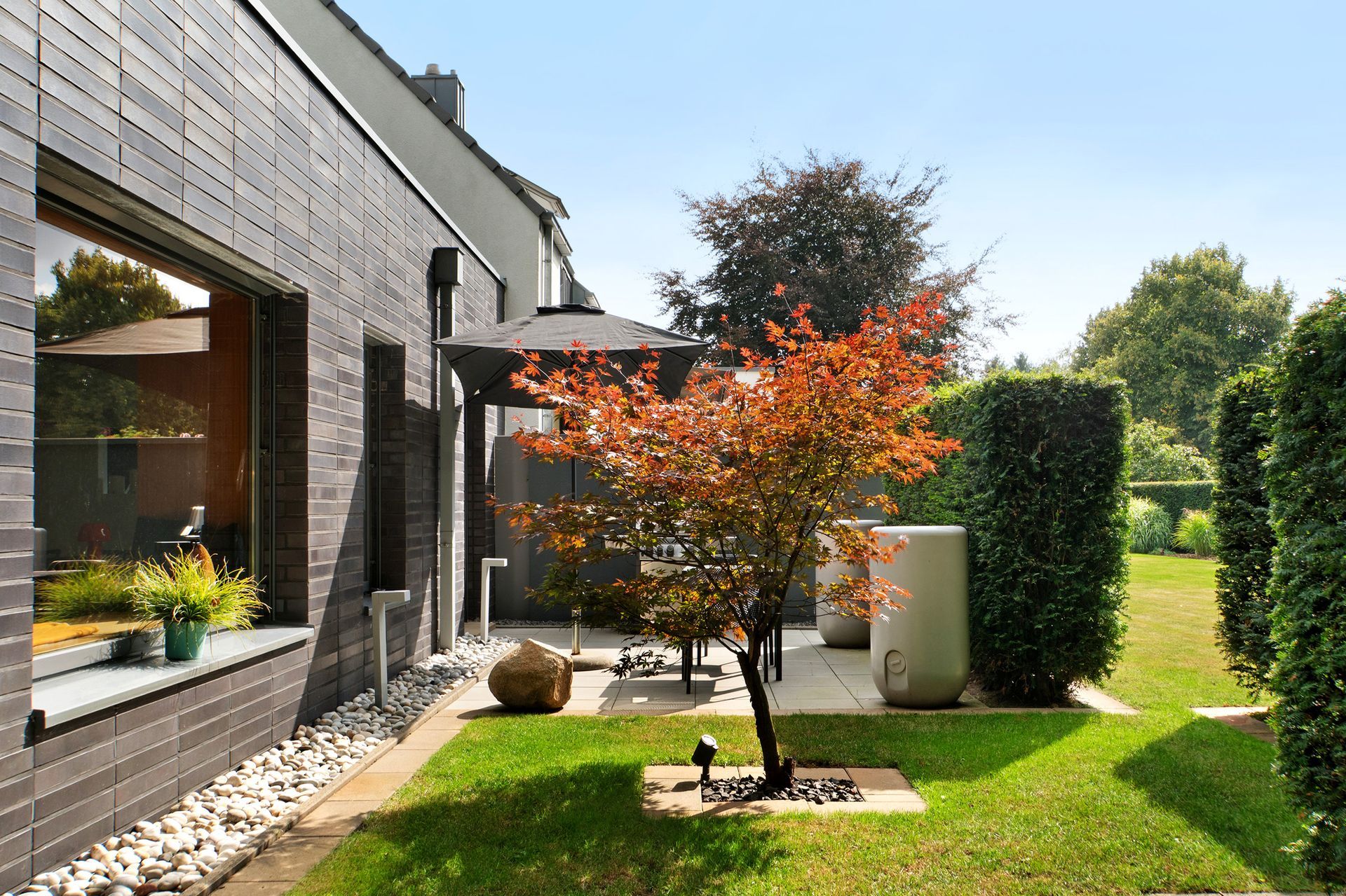 Modernes Haus mit Backsteinfassade, gepflegtem Rasen und einem kleinen Baum mit roten Blättern.