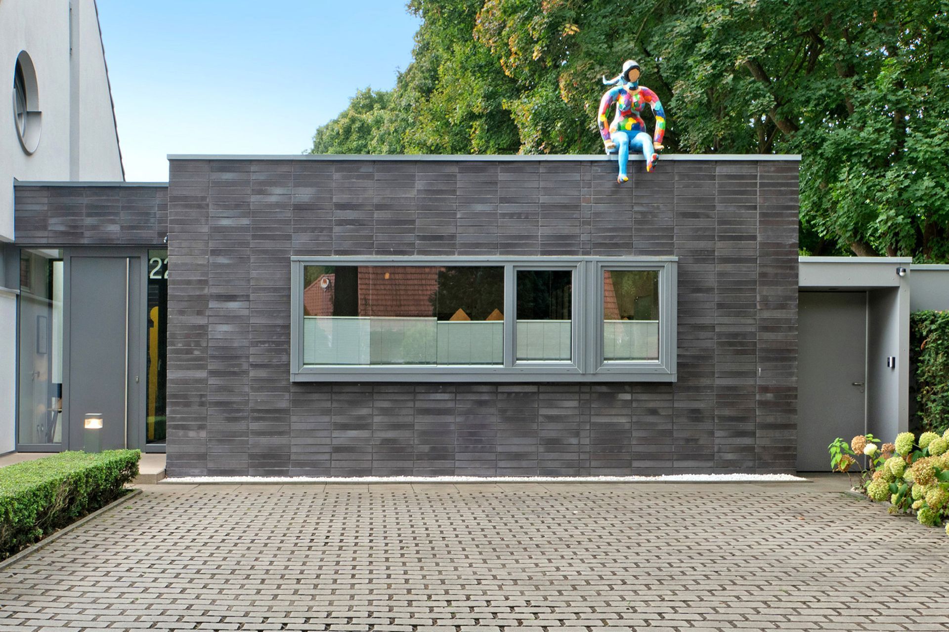 Modernes Gebäude mit grauer Backsteinfassade, Skulptur auf dem Dach und gepflasterter Auffahrt.