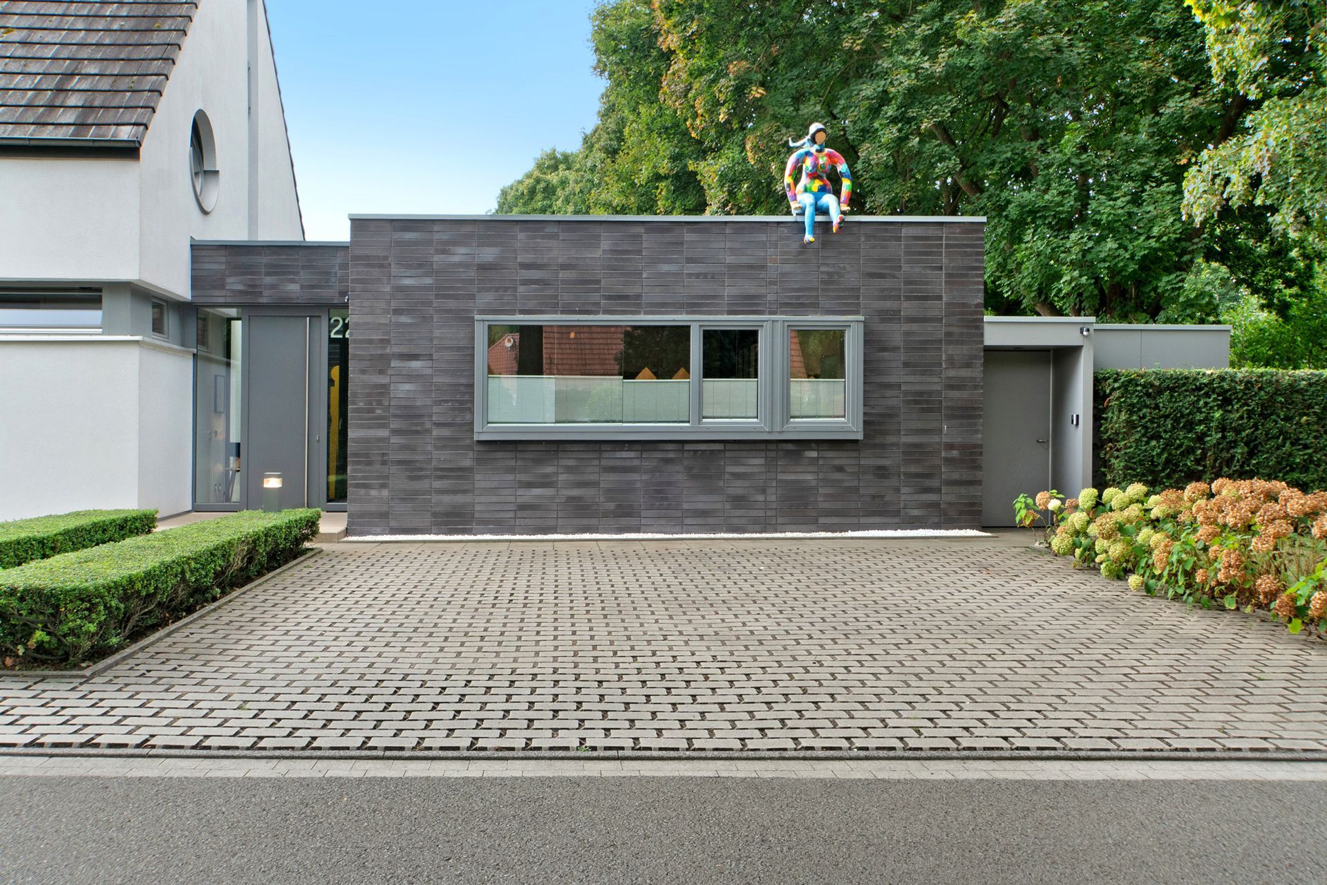 Modernes Haus mit grauer Backsteinfassade, einem Fenster und einer Figur auf dem Dach. Einfahrt mit eingelassenen Objekten.