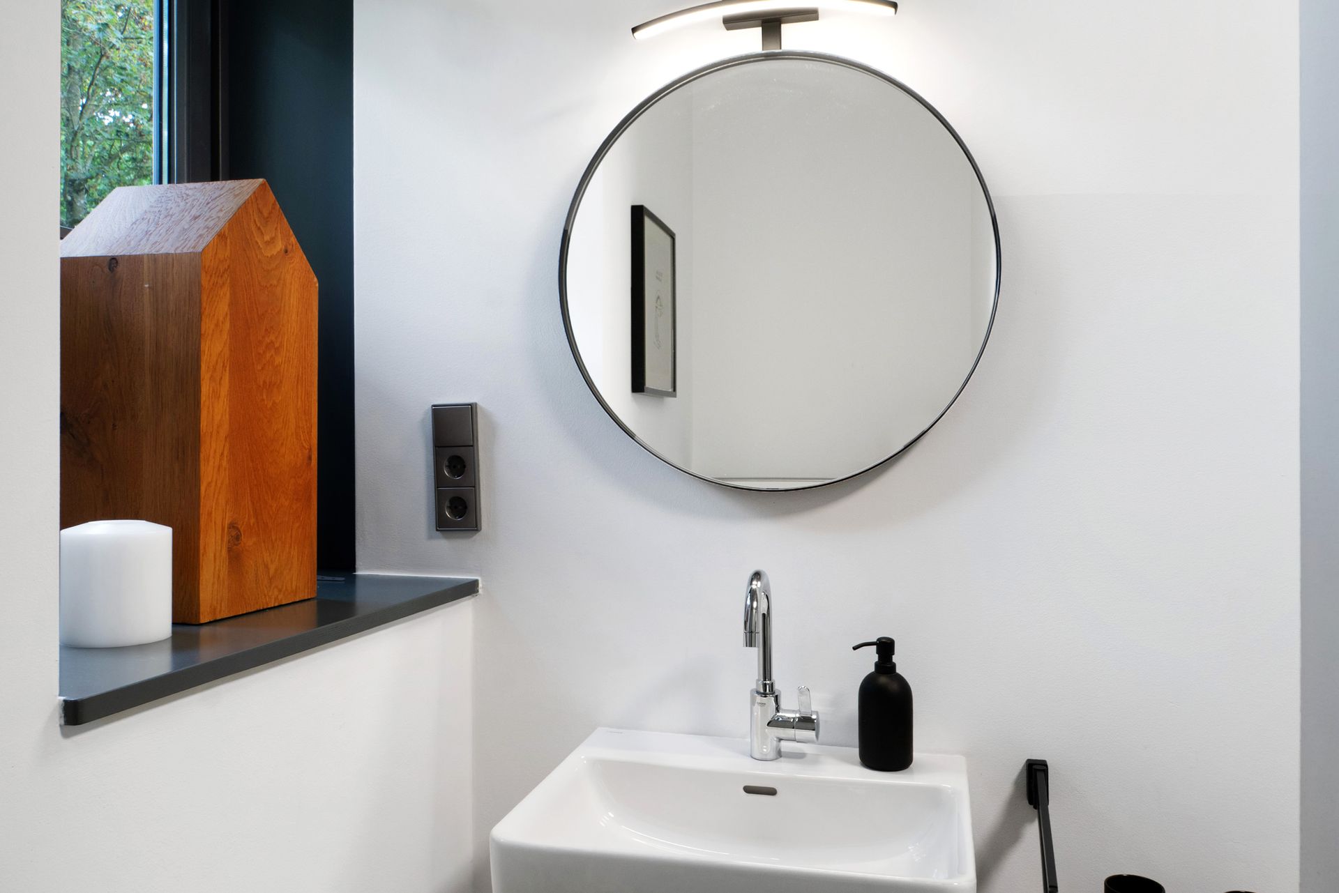 Badezimmer mit rundem Spiegel, weißem Waschbecken, dekorativem Häuschen und schwarzem Seifenspender.