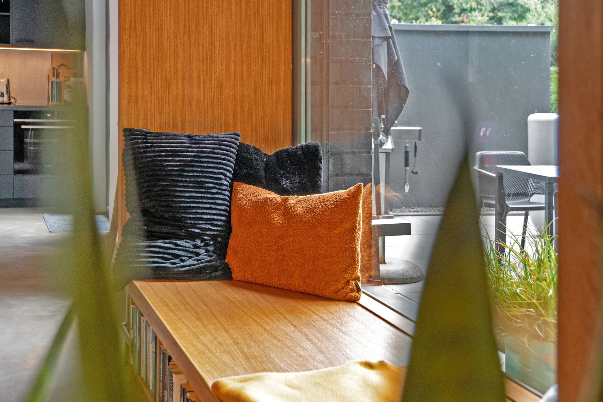Holzbank mit orangefarbenen und schwarzen Kissen, Blick aus dem Fenster auf Terrassenmöbel und Grünanlagen.