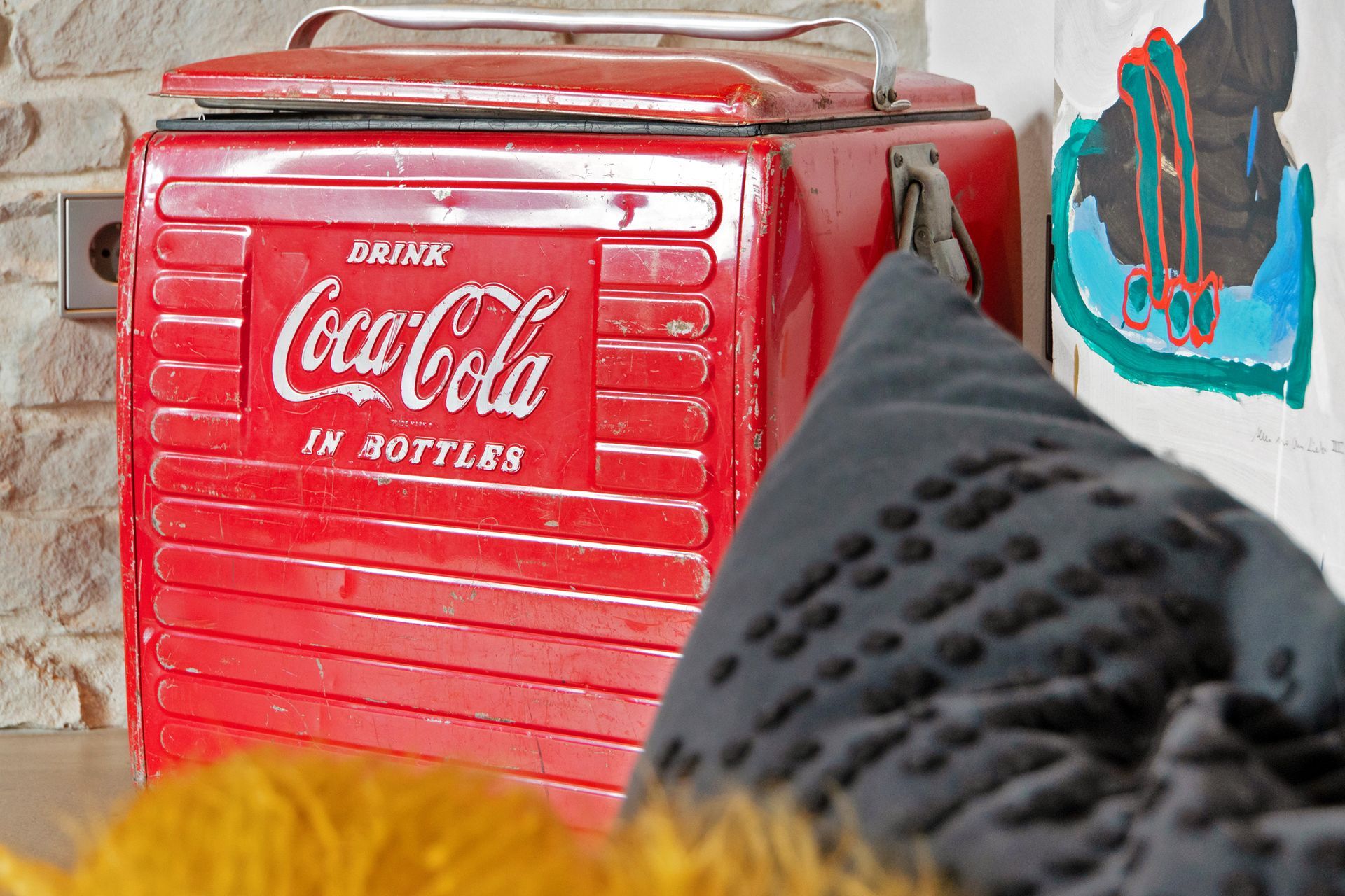 Roter, alter Coca-Cola-Kühler. Graues Kissen im Vordergrund, Kunstwerk an der Wand.
