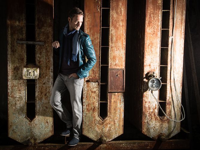 Ein Mann in einer blauen Lederjacke lehnt an einer rostigen Wand.