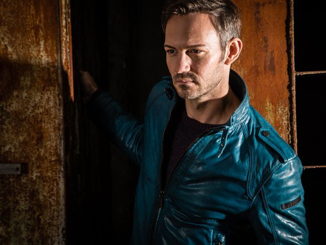 Fashion - FotografieEin Mann in einer blauen Lederjacke steht in einem dunklen Raum.