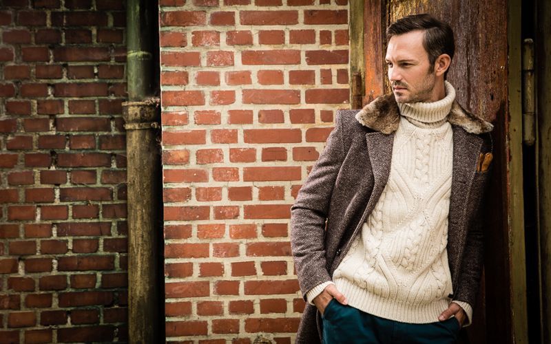 Modefotografie - Ein Mann in Pullover und Mantel steht vor einer Backsteinmauer.