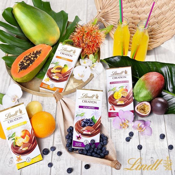 Ein Holztisch mit Schachteln Lindt-Schokolade, umgeben von Früchten und Blumen.