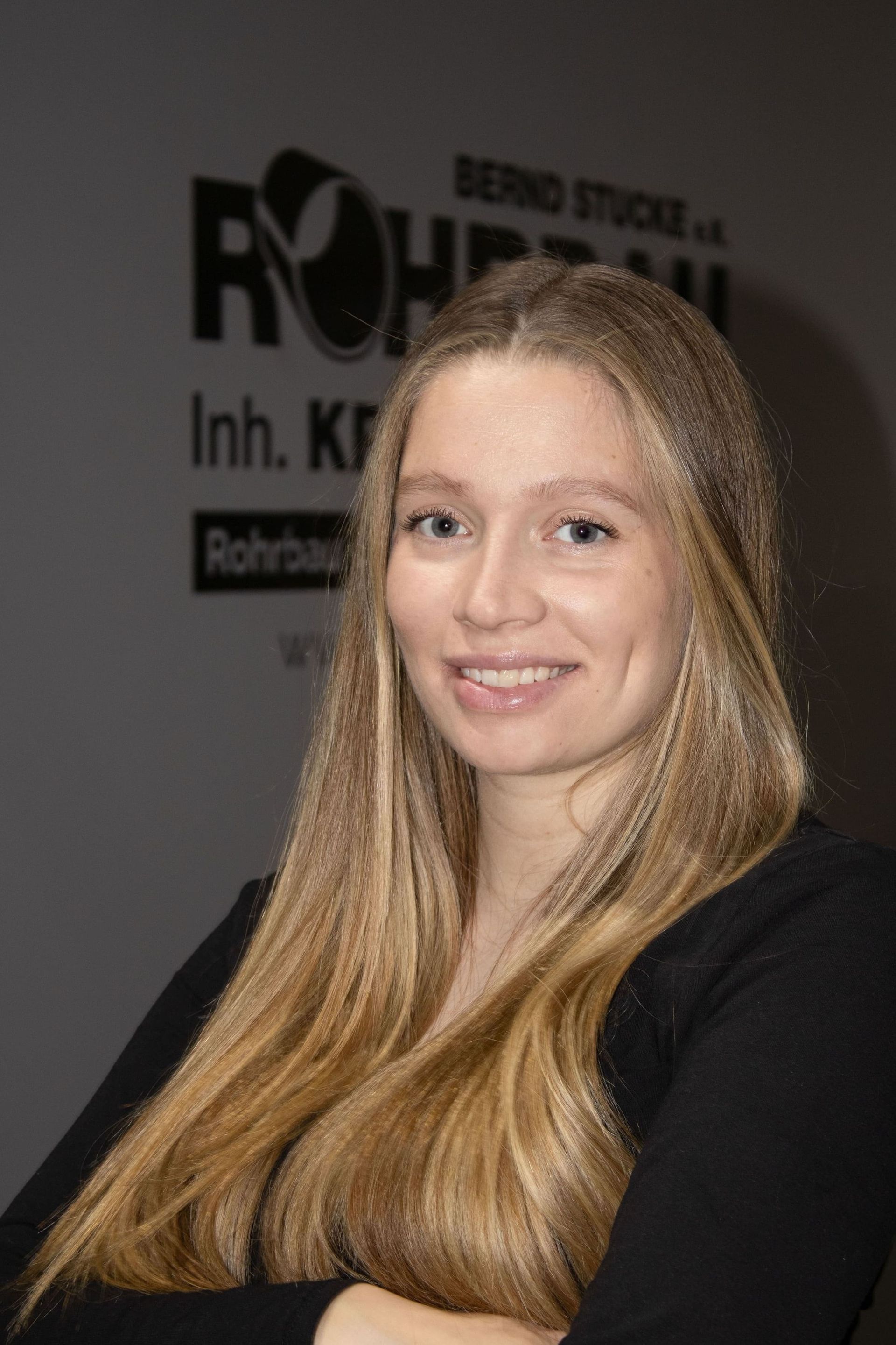 Eine lächelnde Frau mit langen blonden Haaren und schwarzem Oberteil posiert vor dem Hintergrund einer Indoor-Veranstaltung.