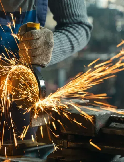 Behandschuhte Hände bearbeiten Metall mit einem Schleifgerät und erzeugen dabei hellorange Funken in einer Werkstatt.