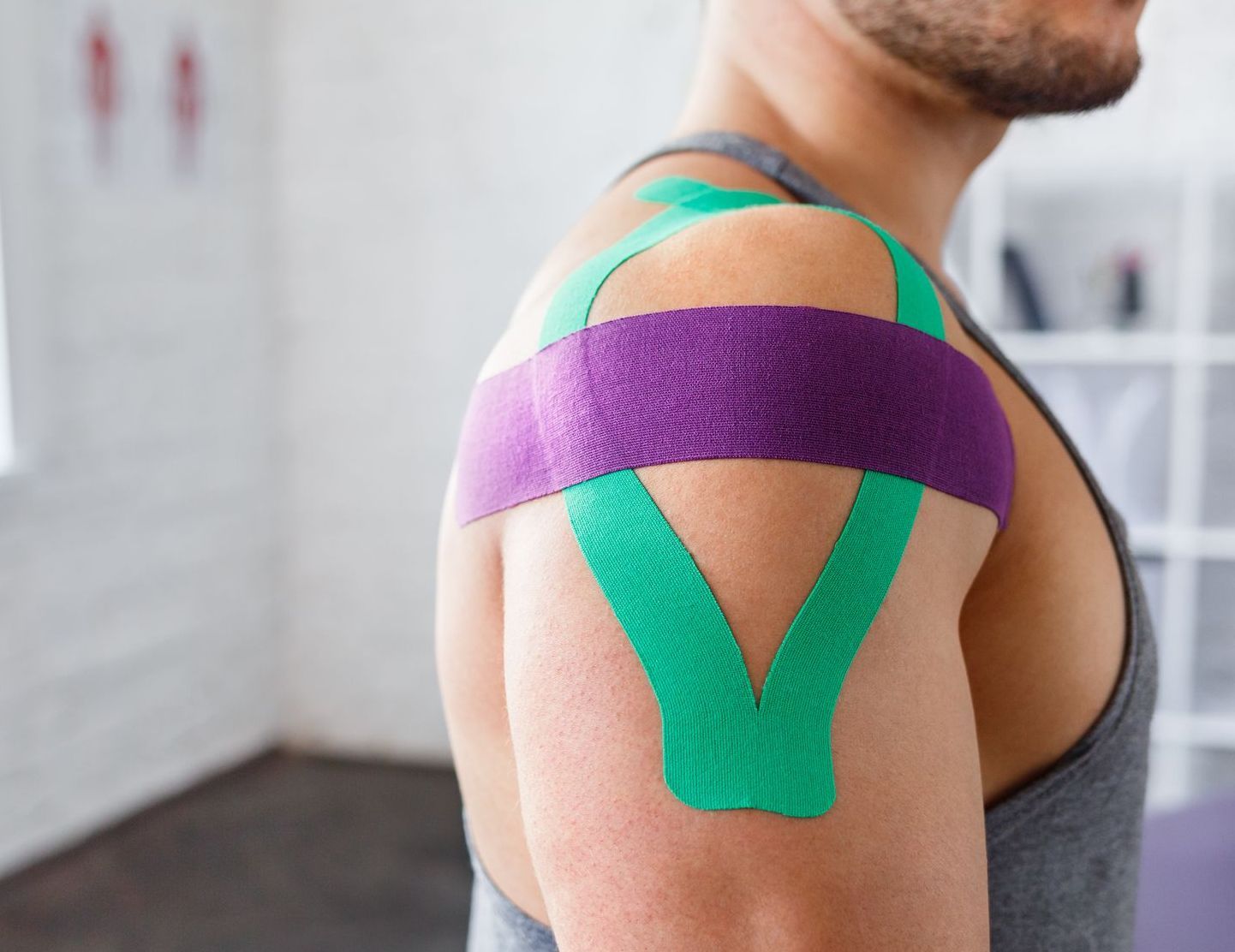 Männerschulter mit lila und grünem Kinesiologie-Tape in einem Fitnessstudio.