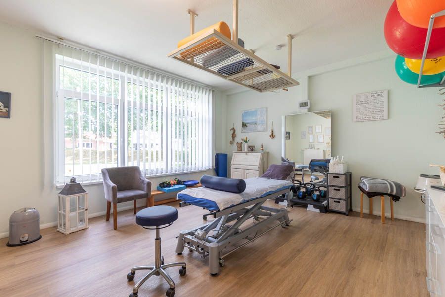 Therapieraum mit Massagetisch, Fenster, Geräten, Stuhl und Hängekonstruktion.