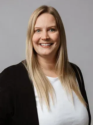 Lächelnde blonde Frau, die ein weißes Hemd und eine schwarze Strickjacke vor grauem Hintergrund trägt.